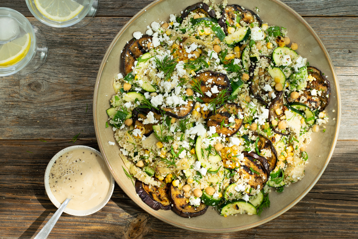 Bovenaanzicht van een couscoussalade met veel groene kruiden en gegrilde aubergine, op een ruwe houten ondergrond.