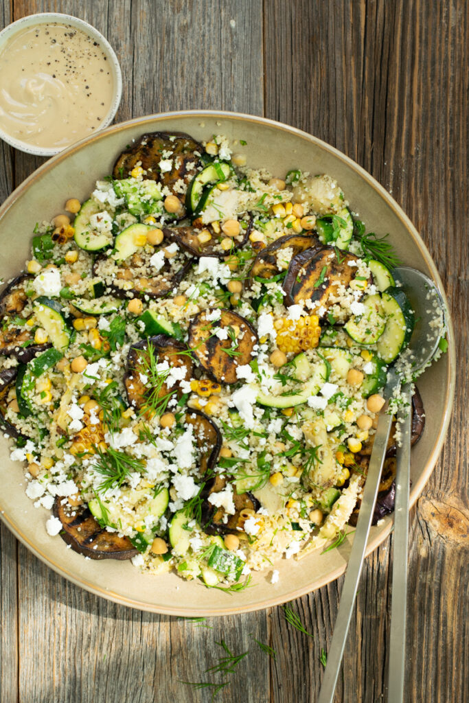 Bovenaanzicht van een couscoussalade met veel groene kruiden en gegrilde aubergine, op een ruwe houten ondergrond.