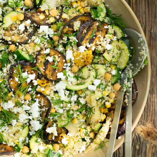 Bovenaanzicht van een couscoussalade met veel groene kruiden en gegrilde aubergine, op een ruwe houten ondergrond.