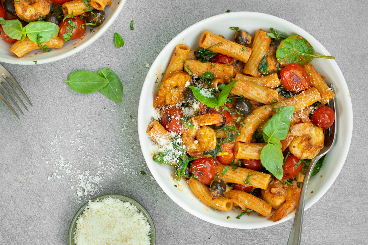 Bovenaanzicht van een diep wit bord met tortiglioni met paprikasaus, kerstomaten, zwarte olijven en scampi's, afgewerkt met basilicumblaadjes en Parmezaanse kaas.