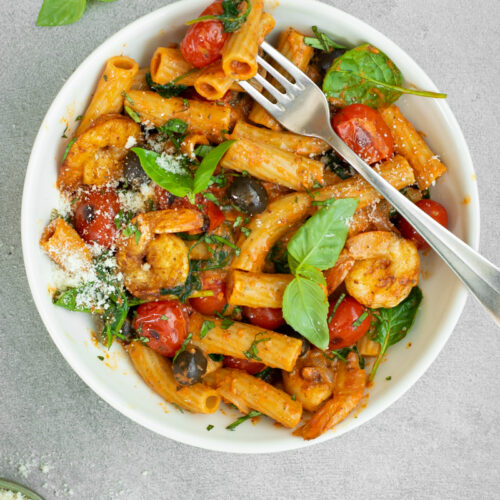 Bovenaanzicht van een diep wit bord met tortiglioni met paprikasaus, kerstomaten, zwarte olijven en scampi's, afgewerkt met basilicumblaadjes en Parmezaanse kaas.