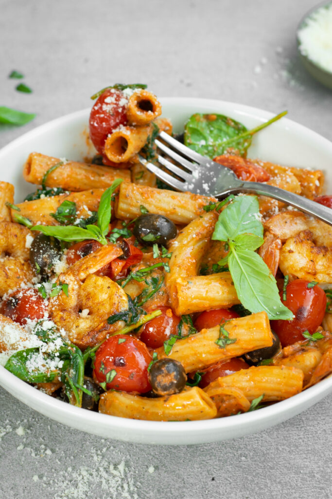 Zijaanzicht van een diep wit bord met tortiglioni met paprikasaus, kerstomaten, zwarte olijven en scampi's, afgewerkt met basilicumblaadjes en Parmezaanse kaas.