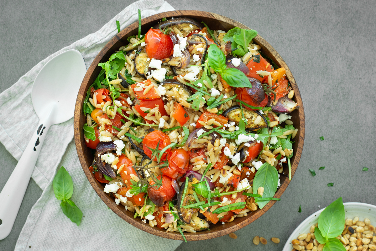 Bovenaanzicht van orzosalade met geroosterde aubergine, paprika en rode ui, afgewerkt met rucola, vers basilicum en feta.