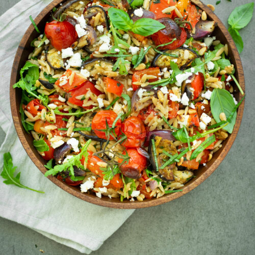 Bovenaanzicht van orzosalade met geroosterde aubergine, paprika en rode ui, afgewerkt met rucola, vers basilicum en feta.